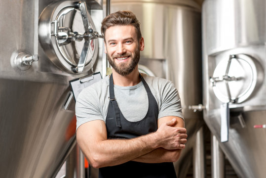 A portrait of handsome brewer in uniform at the beer manufacture with metal containers on the background