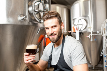 Handsome brewer in uniform tasting beer at the brewery with metal containers on the background