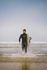 Surfer in wetsuit with his surfboard leaving the sea after surfing. Outdoor beach water sport and surf lifestyle.
