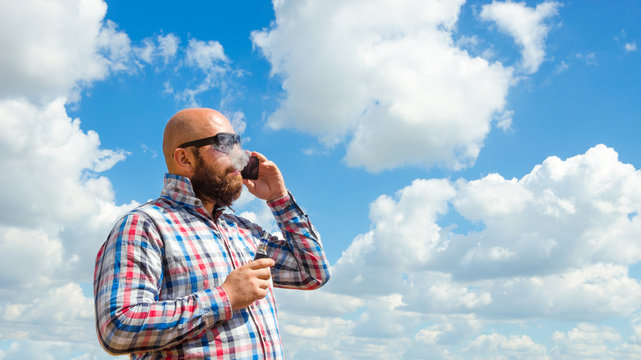 Hairless man with beard in glasses smoke electronic cigarette. outdoor banner blue sky w clouds background.
