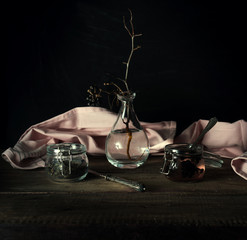 rustic still life. dry twig in a glass vase, pink drape and jars on a wooden table. black background
