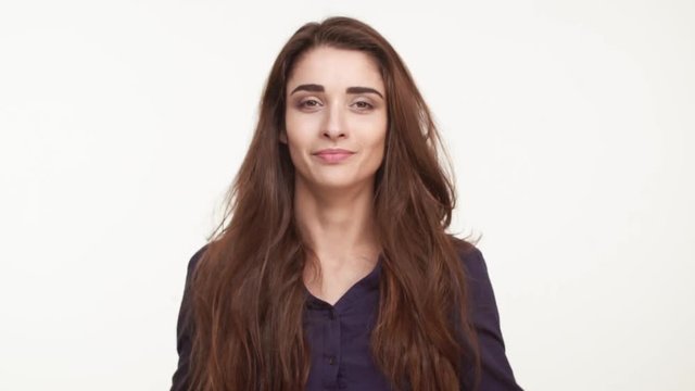 Beautiful Caucasian female in dark blue shirt and with long brown hair standing on white background in amazement keeping head in hands in slowmotion