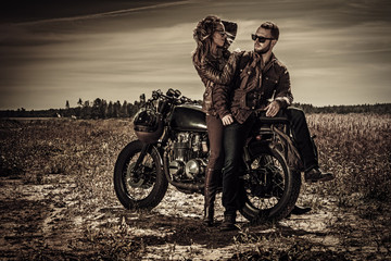 Young, stylish cafe racer couple on vintage custom motorcycles in field - obrazy, fototapety, plakaty