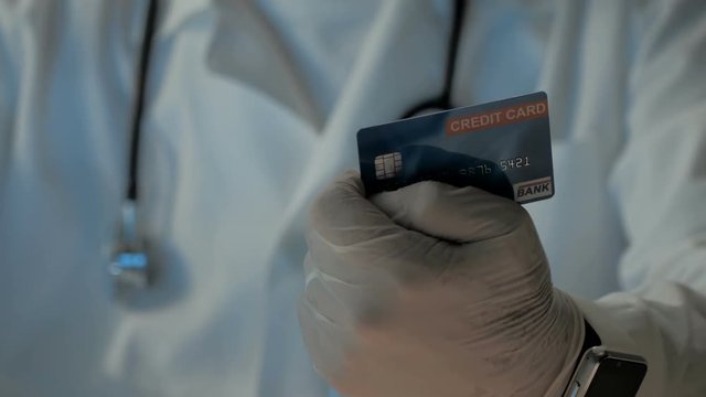 Doctor in white medical gown with stethoscope. Hand in latex gloves holds a credit card. Professional employee gets information data, date expired, name, number, CVC from debit card, turns, twist it