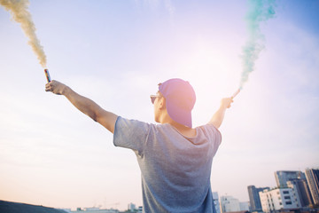 Male teenager holding colorful smoke sticks up in the air over urban city background