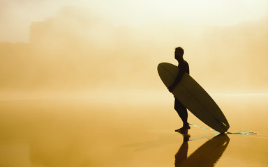 Surfer holding his surfboard and looking for waves on a misty urban beach. Outdoor beach water sport and surf lifestyle.