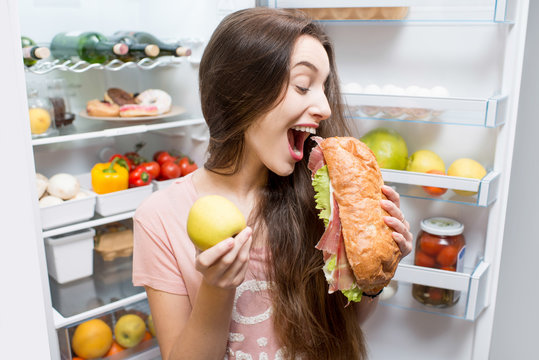 Young woman choosing between apple and big sandwich standing in front of the refrigerator full of vegetables and fruits