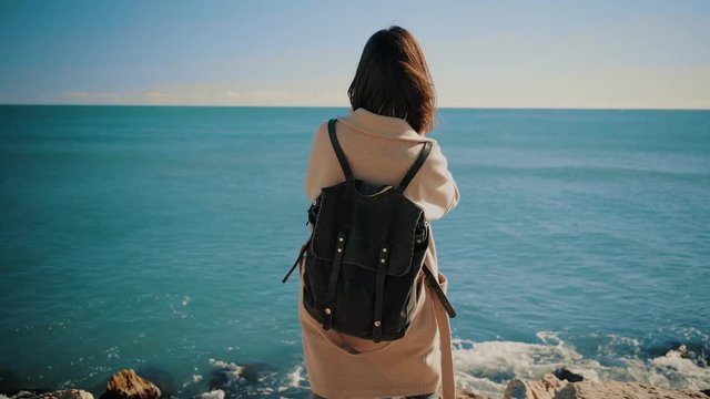 Back view on traveller brunette girl stays on big rocks in front of deep blue see and takes pictures with her smart phone.Camera slowly moves around and stops in front of sun position, overexposed