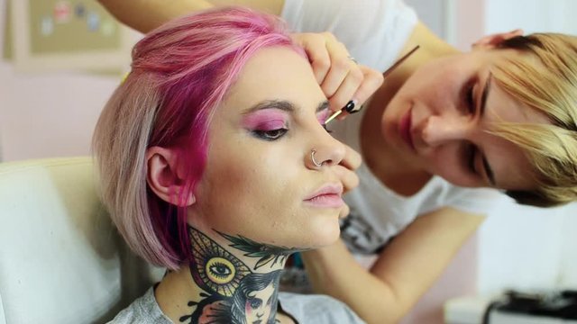 Professional makeup artist applies makeup to a model with tattoos and pink hair.