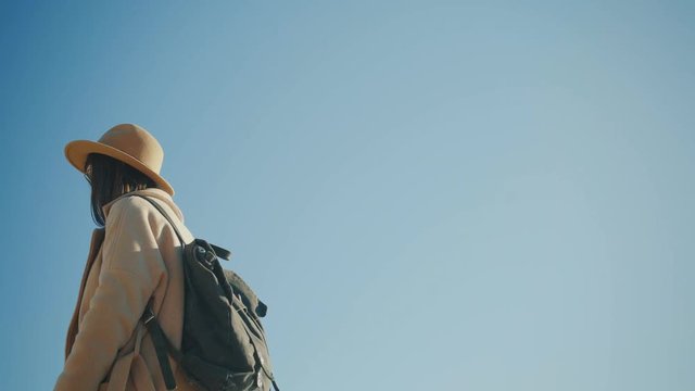 Exited traveller girl in coat with black backpack and in felt hat says wow, looking around and isolated on blue sky at sunny day
