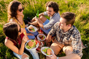 Junge Männer und Frauen sitzen zusammen im Gras und essen Hamburger auf einer Grillparty auf Wiese