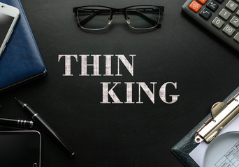 Black chalkboard with business accessories (notepad, magnifying glass, fountain pen, tablet, phone, glasses and calculator) and text THINKING. Top view.