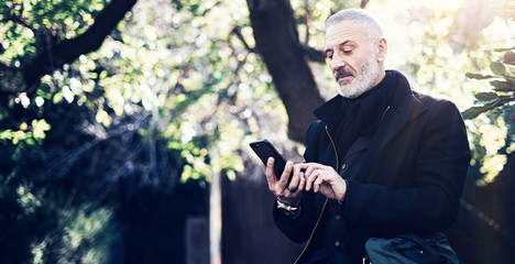 Fototapeta premium Portrait of successful middle age businessman using modern smartphone while spending time in city park at sunny day.Horizontal wide,blurred background.