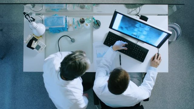 Top View of Two Doctors at the Working Desk Discussing Patient's X-Ray Shown on a Monitor Screen. They Come to Conclusion and Doctors Writes Down Diagnose. Shot on RED EPIC-W 8K Helium Cinema Camera.