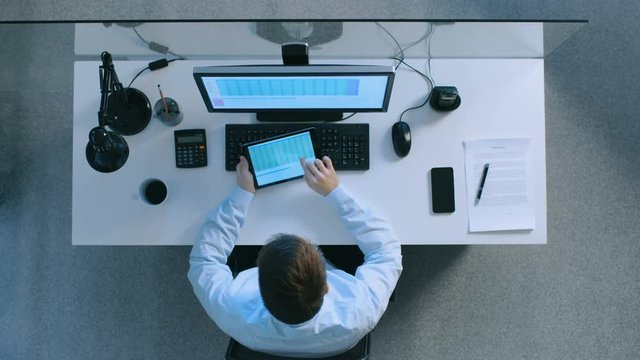  Top View of an Office Worker Sitting at His Desk  Browsing Through Spreadsheets on His Tablet Computer. He Also Uses His Personal Computer. Shot on RED EPIC-W 8K Helium Cinema Camera.
