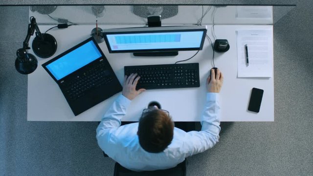 Top View of IT Specialist Working with Spreadsheets and Documents on His Laptop and Personal Computer Simultaneously. Stack of Signed Papers on His Desk. Shot on RED EPIC-W 8K Helium Cinema Camera.