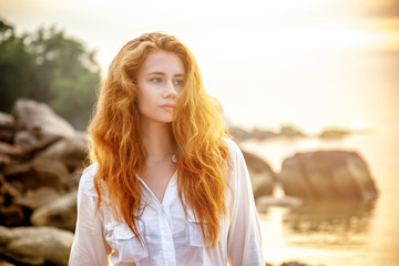 Beautiful young woman with long red hair on the beach at sunset