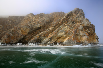 Fototapeta na wymiar Rocky shore of lake Baikal in the fog in winter Rocky headland on the Western coast of lake Baikal sticks in ice-covered body of water, Fog hampers visibility
