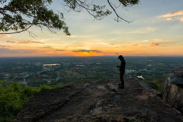 Man traveler is using digital phone on cliff with beautiful landscape sunset over cliff and city  - success concept