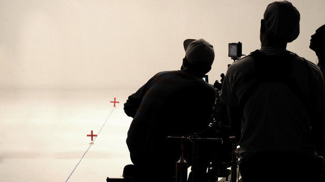 Behind the scenes of silhouette film crew team are setting camera and making technique for chroma keys on commercial movie with high quality equipments such as tripod monitors and red mark on floor.