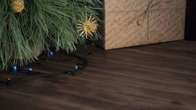 The man puts box of gift under the Christmas tree. Fir stand on the wooden table and decorated with glowing garland and Christmas toys. Box with gift wrapped in brown paper and tied with twine.