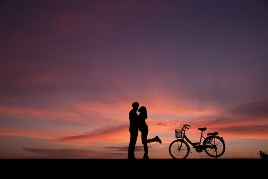 Silhouette of lovers standing hug and kiss with vintage bikes on the side.The background image is a sunset in Thailand.