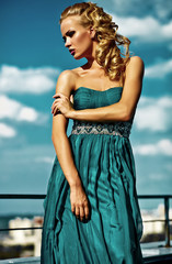 Young sexy blond woman model  in evening dress posing on blue sky background