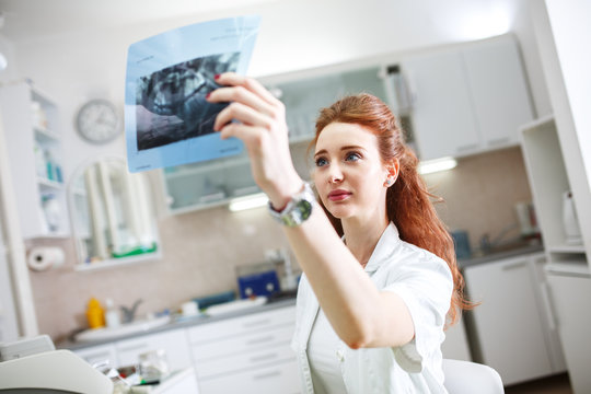 Female red hair dentist in dental office examining x-ray image.