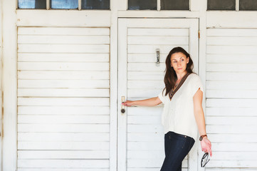Fototapeta premium Outdoor fashion portrait of 35-40 year old woman wearing white marine pullover and dark denim jeans, holding sunglasses, standing against white wooden background