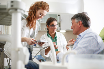 Dentist and her female assistant in dental office talking with senior patient and preparing for...