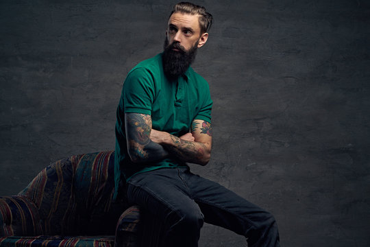 Tattooed bearded male sits on a chair.