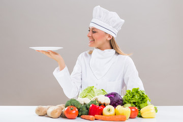 Female chef surrounded with vegetables holding plate.
