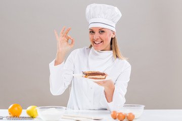 Beautiful female confectioner is showing ok sign while holding slice of cake.