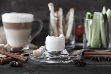 Soft Boiled egg with small spoon, coffee latte, sugar slices, bread for breakfast on old dark grey rustic wooden table. Healthy food. Retro newspaper and cinnamon for decoration. Low key dark photo.