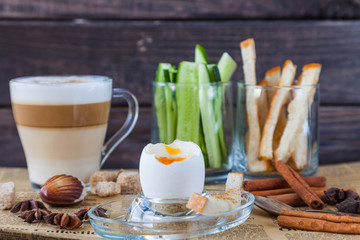 Obraz na płótnie Canvas Soft Boiled egg with small spoon, coffee latte, sugar slices, bread for breakfast on old dark grey rustic wooden table. Healthy food. Retro newspaper and cinnamon for decoration. Low key dark photo. 