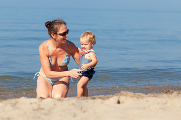 Happy pretty baby girl and mather in swimsuit playing with sand on beach