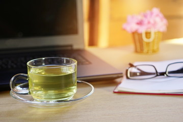  an office desk on which there are various objects : a cup of tea, a laptop, 
 a notebook, flower pot and glasses.