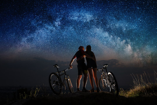 Rear view man and woman are hugging, standing on a hill with mountain bicycles under the starry sky and looking at each other. Night landscape with colorful Milky Way