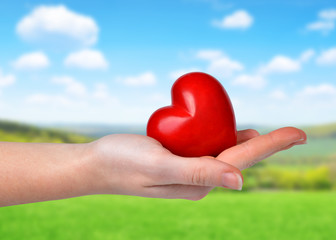 Naklejka na ściany i meble Hand holding red heart on the background blurry landscape with blue sky. 