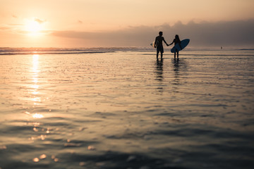 Silhouettes of a couple holding surf boards in hands walking at sunset on coastline