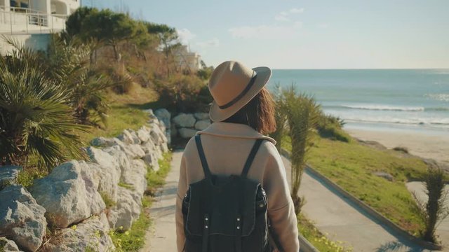 Camera follows behind attractive brunette girl in fedora hat and wool coat on walking down to sand beach next to sea through small narrow lane with green palms at sunny day.