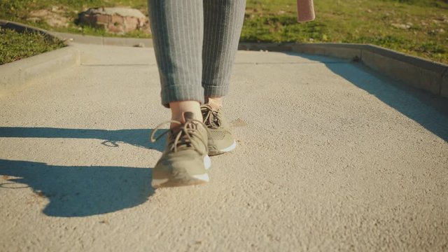Camera follows front woman legs in sneakers walking down through small narrow lane with greenery on sides at sunny day.