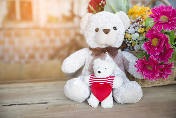 Two teddy bears sitting on a wooden table,little bear sit hugging red heart In the background is a bouquet of flowers