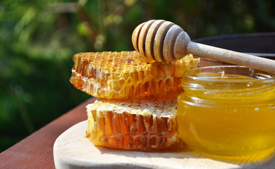 Pieces of honeycomb with spoon and glass jar with honey are on a wooden board. Outdoor background.