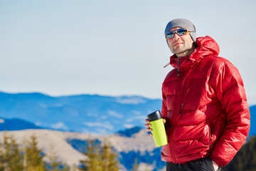 hiker in red down jacket with big plastic cup resting near the tent in the Carpathians mountains at winter