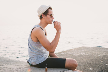 Young hipster man sitting and vaping on sea front