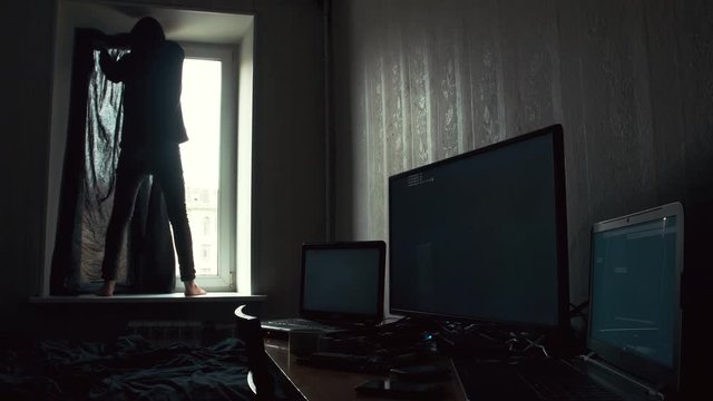 Young in dark hood standing on the windowsill and covering windows with black curtain