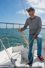A man in a striped sweater, jeans, a blue cap and red sneakers controls sailing boat. Boy holds the...