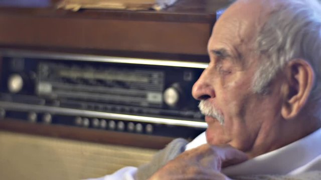 Old Man is Looking up and Listening to Radiogram Drinks Tea Sitting in Cosy Living Room and Resting at Home in Cold Wintry Day at Home Wrinkled Face of Man