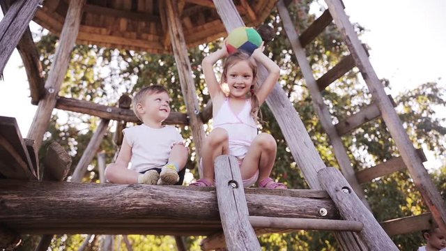 Beautiful smiling girl and boy siblings playing with ball outdoor on tree house together with their parent on sunny day
